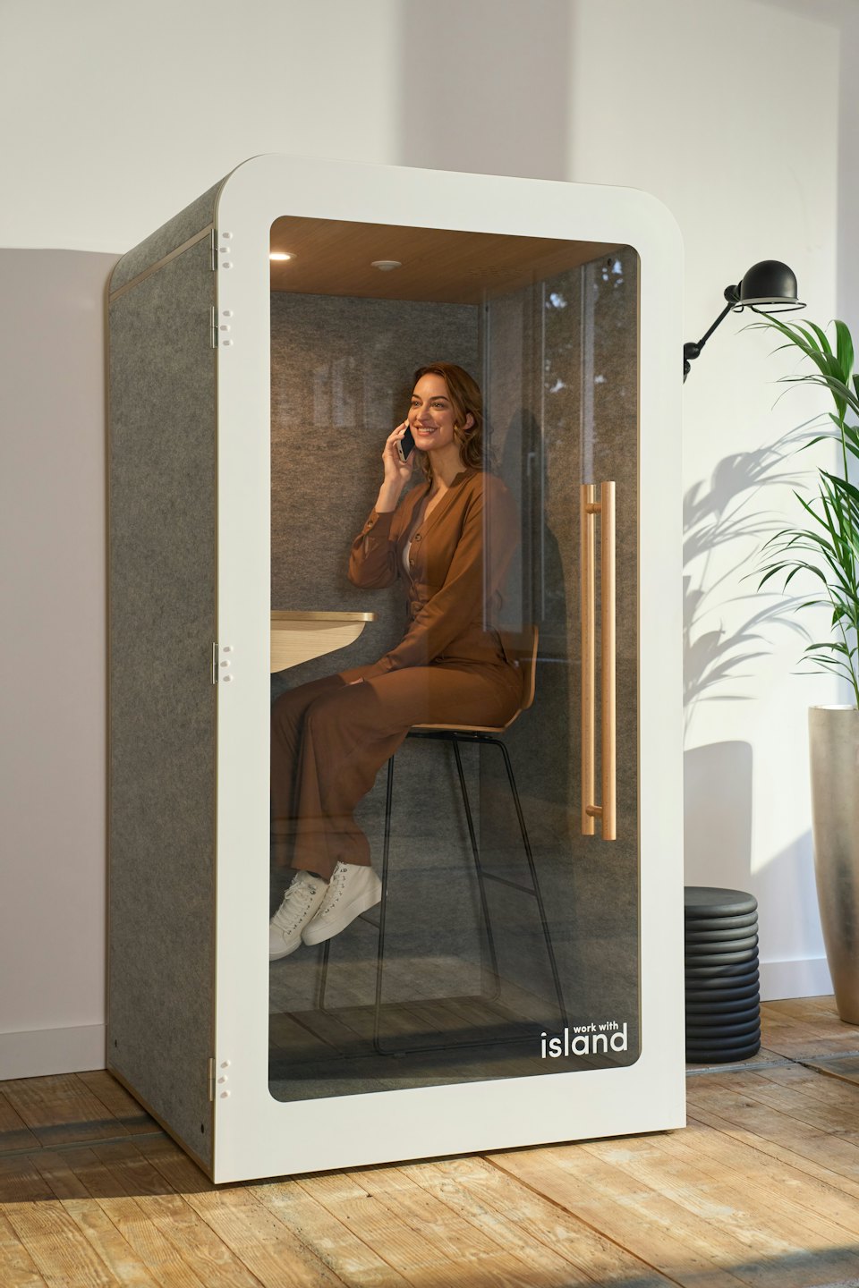 Žena v hnědé kombinéze mluvící po telefonu v kancelářském boxu.